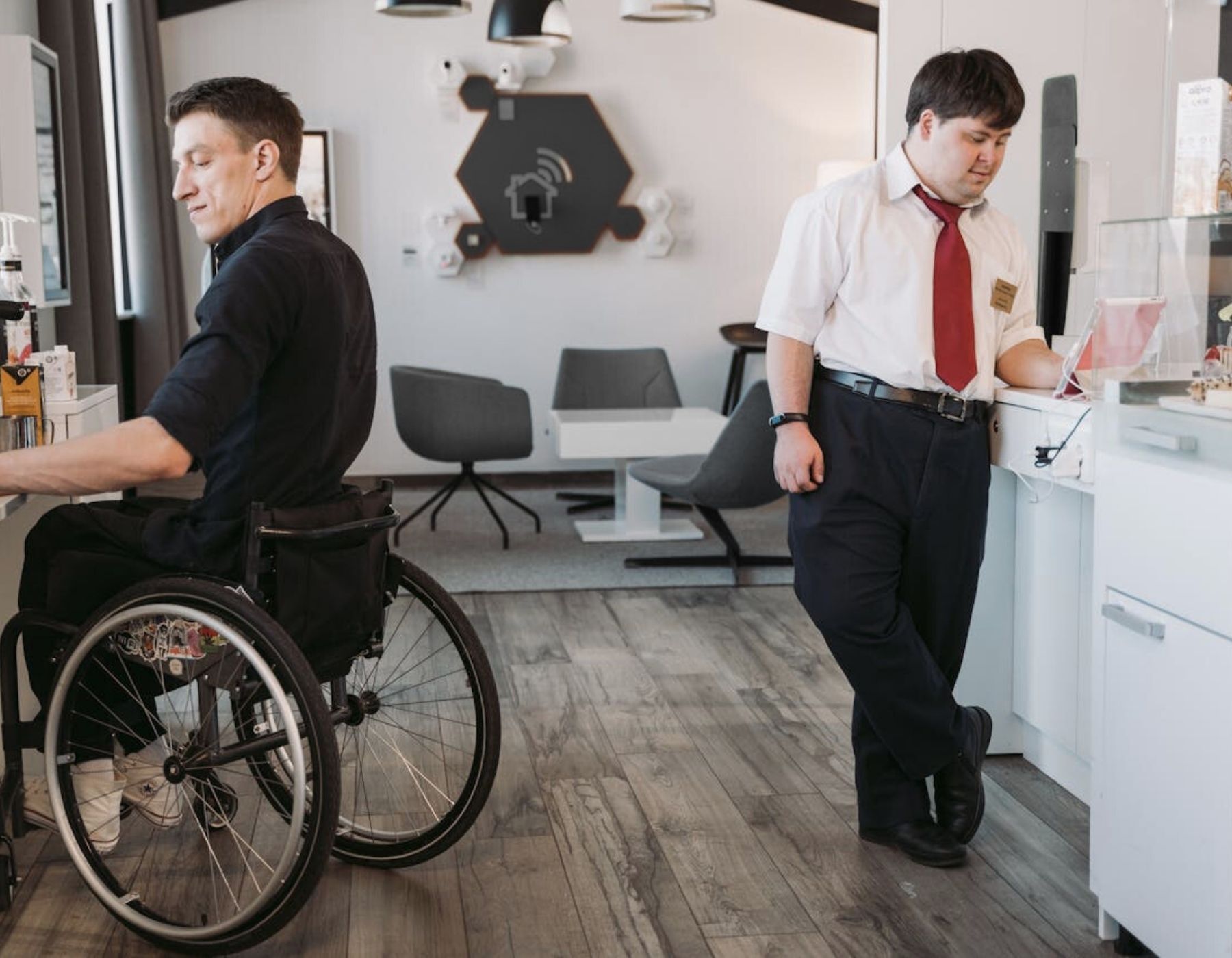 Foto zweier Männer mit einer Behinderung. Ein Mann sitzt im Rollstuhl und bereitet sich gerade einen Kaffee zu. Der andere Mann arbeitet am Tablet.