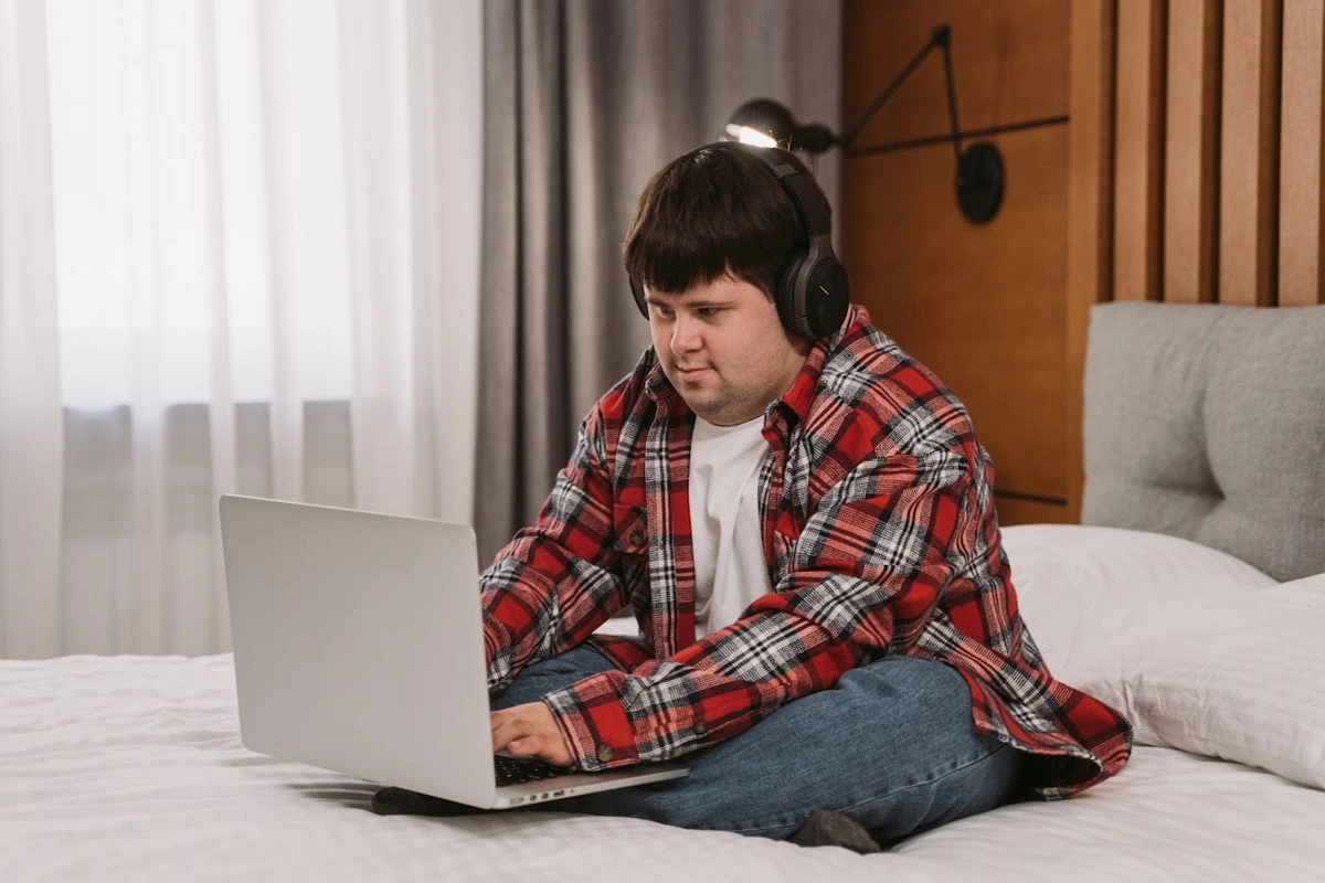 Ein Mann mit einer Behinderung arbeitet am Laptop. Er sitzt auf einem Bett und trägt Kopfhörer.